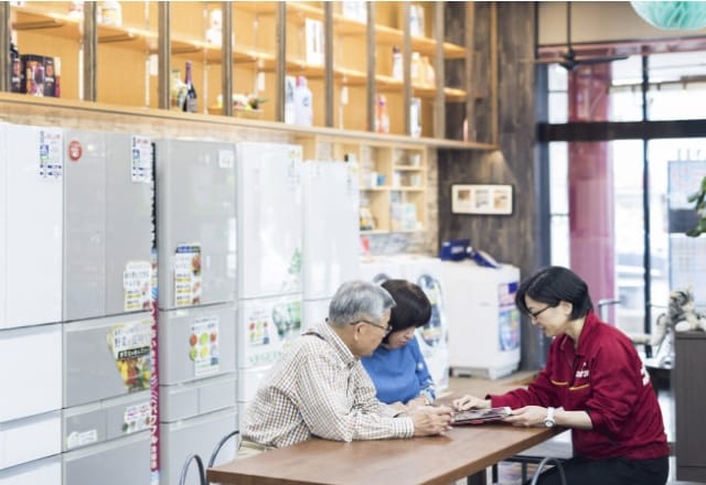 写真:店内のテーブルでスタッフに相談しているひと。背景には冷蔵庫や洗濯機が並んでいる。